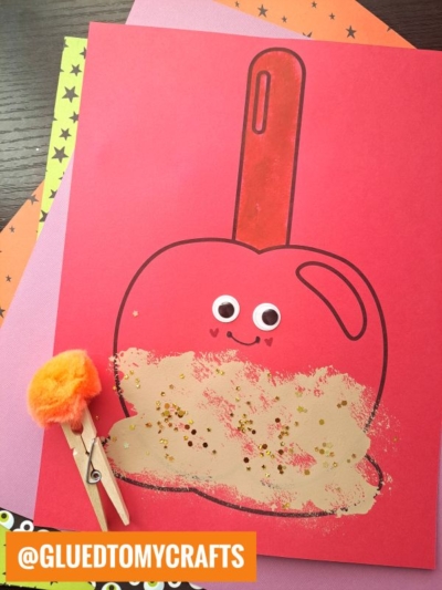 A red paper with a stamped caramel apple drawing, googly eyes, and glitter. An orange pom-pom is clipped to a clothespin beside the paper. Decorative colored sheets are underneath. Text reads @GLUEDTOMYCRAFTS.
