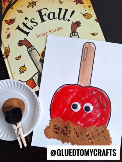 A children's book titled Its Fall! lies next to a craft project: a stamped caramel apple with googly eyes, brown paint, and sprinkles, plus a clothespin-painted sponge and a brown paint puddle on a paper plate.