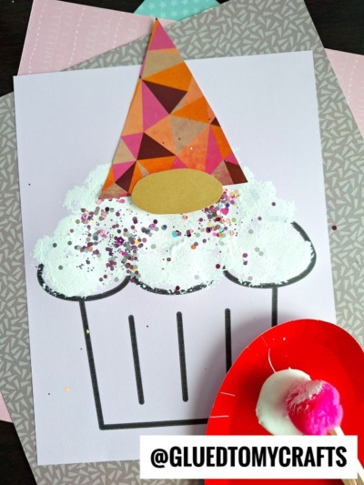 A gnome cupcake craft made with white paint, colorful glitter, and patterned paper on a sheet. A small red plate with white paint and a pom-pom is nearby. The image has the text @GLUEDTOMYCRAFTS at the bottom.