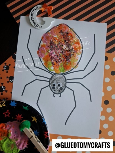 A Halloween spider craft made with orange and green painted fingerprints for the body, black marker legs, and a drawn spider face on white paper. A pom pom stamped spider adds extra texture, while decorative papers and paint supplies are placed around.