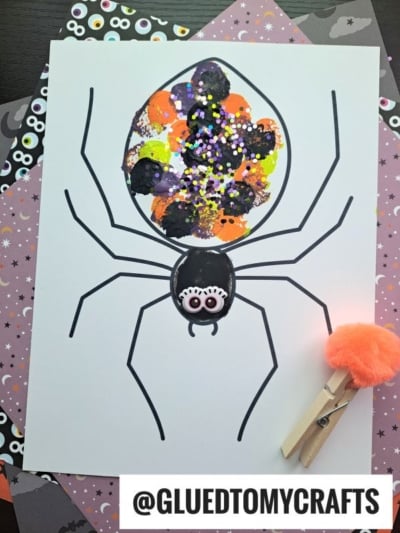 A pom pom stamped spider drawing with a body made of colorful paint blobs and glitter. A decorated clothespin with orange fluff sits on the side. Patterned paper with eyes is underneath. Text reads @GLUEDTOMYCRAFTS.