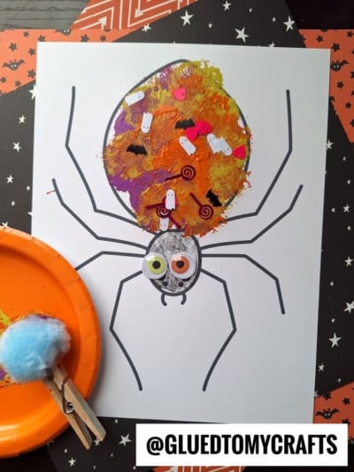 A pom pom stamped spider coloring page features a painted, decorated body covered in Halloween confetti on a black star-patterned background. A blue pom-pom on an orange plate sits nearby. @GLUEDTOMYCRAFTS is written at the bottom.