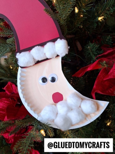 A Santa face Christmas craft inspired by the crescent moon Santa, made from a paper plate, red and pink paper, cotton balls for the beard and hat trim, and googly eyes. @GLUEDTOMYCRAFTS is at the bottom, hanging on a Christmas tree.