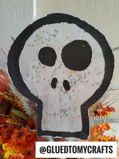 A paper skull craft with black eyes and nose, covered in star glitter, is displayed among orange flowers. Reminiscent of a stained glass skull, this charming decoration adds a touch of whimsy to any space. The Instagram handle @GLUEDTOMYCRAFTS appears at the bottom.