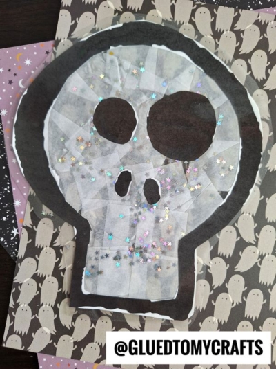 A stained glass skull craft made of black and white paper, tissue, and glitter stars sits on ghost-patterned paper. @gluedtomycrafts is written at the bottom.