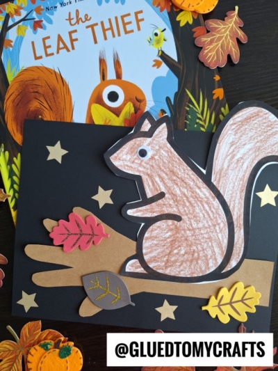 A hand-colored squirrel on tree branch craft sits amid colorful paper leaves on a black background, placed atop the book The Leaf Thief and surrounded by autumn-themed decorations. @GLUEDTOMYCRAFTS.