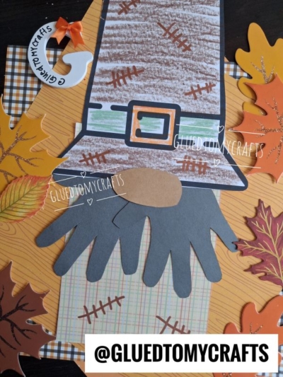 A handprint scarecrow gnome craft made with paper, surrounded by paper leaves. A tag and @GLUEDTOMYCRAFTS watermark are visible on the photo. The scarecrow gnome features a hat, handprint arms, and a brown oval face.