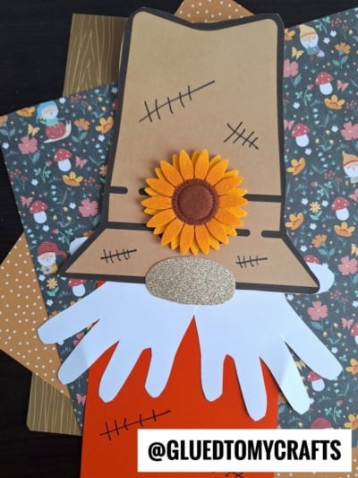 A handmade scarecrow gnome craft made from paper, featuring a large sunflower, a gold glitter nose, white paper handprint beard, and accented with orange, tan, and patterned backgrounds. @GLUEDTOMYCRAFTS is labeled at the bottom.