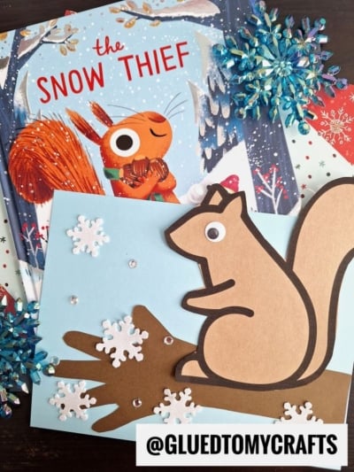 A children's book titled The Snow Thief featuring a squirrel is placed next to a squirrel on tree branch craft on blue paper with snowflakes and sequins, surrounded by decorative snowflakes. The @GLUEDTOMYCRAFTS handle is visible.