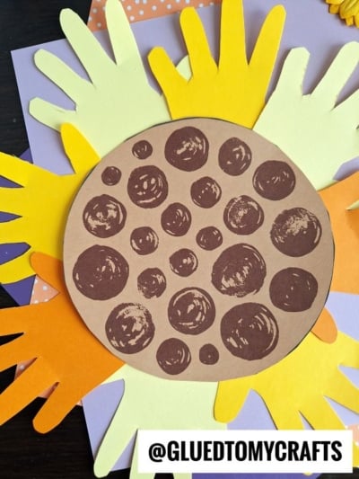 A craft project of a handprint sunflower made from layered yellow and orange paper handprints, with a brown circle featuring darker spots in the center, displayed on a light background. Text reads @GLUEDTOMYCRAFTS.