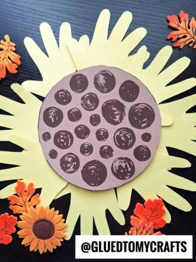 A handprint sunflower craft project with a brown, spotted paper center, yellow paper petals, and accents of artificial autumn leaves and flowers. @GLUEDTOMYCRAFTS is written at the bottom.