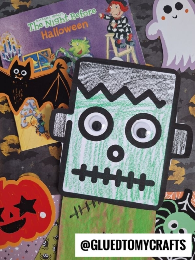 A handmade paper bag Frankenstein craft with googly eyes is surrounded by Halloween decorations, including a bat, ghost, jack-o-lantern, and the book The Night Before Halloween. @GLUEDTOMYCRAFTS text is visible.