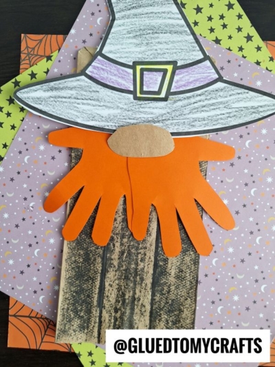 A Halloween craft featuring a lunch sack witch or paper bag witch, with a witch's hat, orange hand-shaped beard, brown nose, and colorful patterned papers on a wood background. Text reads @GLUEDTOMYCRAFTS.