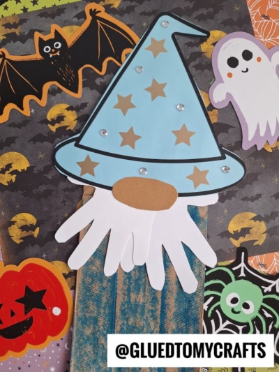A handmade paper bag wizard gnome craft with a blue hat is surrounded by Halloween-themed cutouts, including a bat, ghost, jack-o-lantern, and spider, on a patterned background. @GLUEDTOMYCRAFTS is written at the bottom.