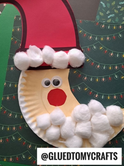 A Santa Claus face crafted from a paper plate, with cotton ball beard and hat trim, googly eyes, and a red paper nose, is set against a festive Christmas background—perfect for adding a whimsical crescent moon Santa touch. @GLUEDTOMYCRAFTS is written below.