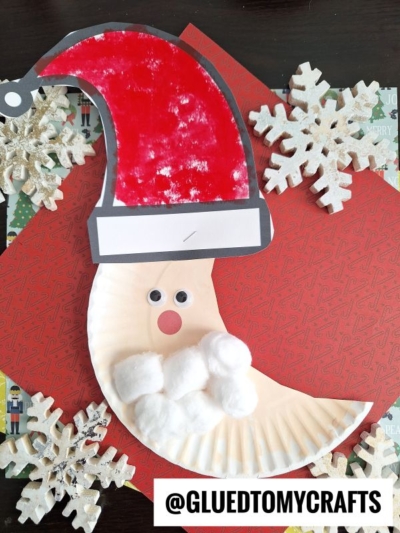 A Santa face craft made from a paper plate, featuring a crescent moon Santa with cotton ball beard, googly eyes, and a red hat. Placed on patterned paper with snowflakes. @GLUEDTOMYCRAFTS is at the bottom.