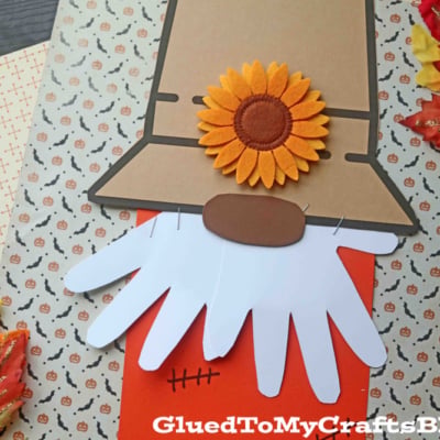 This handmade scarecrow gnome craft features a tan hat with a sunflower, a white beard made from paper hand shapes, and an orange body with stitched details. Decorative autumn leaves and patterned papers complete this charming gnome scarecrow scene.