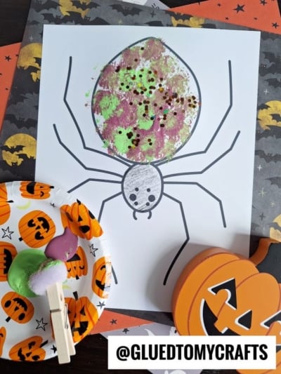 A Halloween-themed pom pom stamped spider coloring page featuring a large spider decorated with green, purple, and black paint and glitter. Nearby are a paint palette, a Halloween napkin, and a wooden pumpkin decoration.