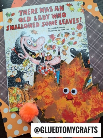 A children’s book titled There Was an Old Lady Who Swallowed Some Leaves! is displayed with a pom pom stamped leaf craft featuring googly eyes and an orange pom-pom on a polka dot and plaid background.
