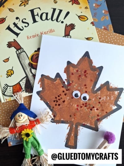 A fall-themed craft scene with a “It’s Fall!” book, a pom pom stamped leaf art project with glitter and googly eyes on white paper, a small scarecrow doll, and a purple dot marker. Text reads @GLUEDTOMYCRAFTS.