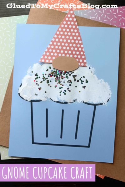 A handmade card features a charming gnome cupcake, with a pink polka dot hat, brown oval nose, white textured frosting, and glitter sprinkles, set against a blue background.
