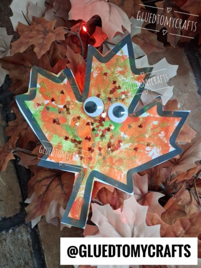 A colorful pom pom stamped leaf craft with googly eyes sits on autumn leaves. The maple leaf is decorated with paint, glitter, and playful pom pom textures. @GLUEDTOMYCRAFTS is written at the bottom and top of the image.