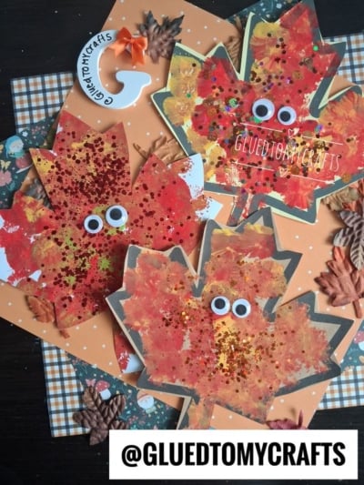 Three autumn leaf crafts, including a charming pom pom stamped leaf decorated with red and gold glitter, googly eyes, and painted textures, are arranged on a patterned background with artificial leaves. The handle @GLUEDTOMYCRAFTS appears twice.