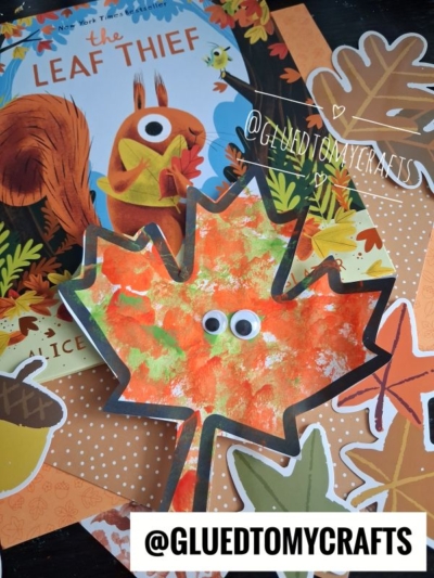 Childrens craft project featuring a maple leaf cutout with googly eyes, autumn-themed paper, and the book The Leaf Thief in the background. The design is completed with a cheerful pom pom stamped leaf for extra texture and fun. @GLUEDTOMYCRAFTS is written at the bottom.