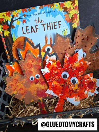 A children’s book titled The Leaf Thief is displayed in a basket with handmade autumn leaf crafts, including pom pom stamped leaf designs decorated with googly eyes, paint, and glitter. Straw fills the basket. The handle @GLUEDTOMYCRAFTS is shown at the bottom.