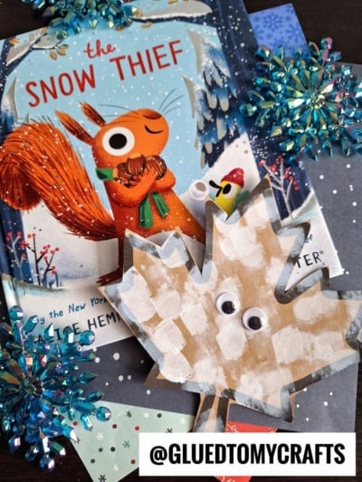 A children’s book titled “The Snow Thief” featuring an illustrated squirrel surrounded by blue snowflake ornaments and a pom pom stamped leaf craft with googly eyes. A label reads @GLUEDTOMYCRAFTS.