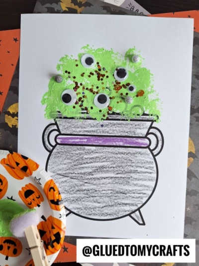 A Halloween craft shows a pom pom stamped cauldron filled with green paint, googly eyes, sequins, and glitter on white paper. Craft supplies and a plate with green paint are nearby, with @GLUEDTOMYCRAFTS written at the bottom.