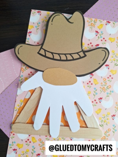 A handmade craft featuring a popsicle stick cowboy hat, a white paper handprint, and a triangle made of popsicle sticks, displayed on floral and chicken-patterned paper. A label reads @GLUEDTOMYCRAFTS.