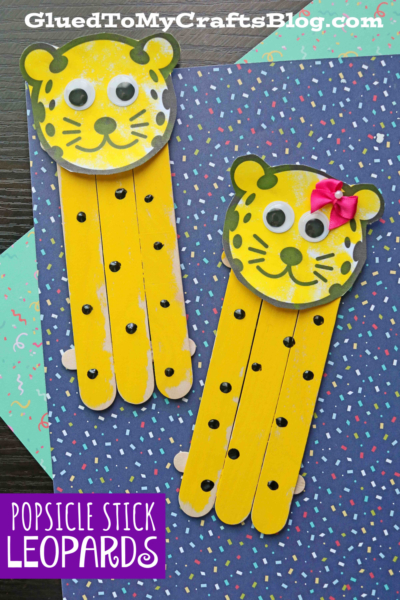 Two yellow popsicle stick leopards with black spots, ears, and drawn faces—one sporting a pink bow—are crafted from paper and sticks. Both are displayed on confetti-patterned paper under the caption Popsicle Stick Leopards.