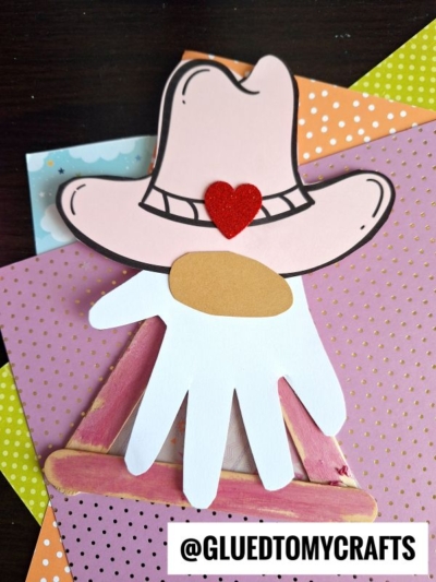 A handmade cowboy gnome craft with a pink hat, red heart, and white hand cutout, placed over colorful polka dot papers. The handle @GLUEDTOMYCRAFTS appears at the bottom.