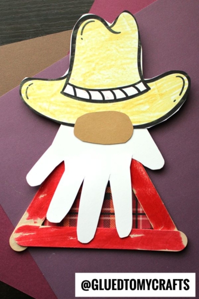 A cowboy gnome craft made with a yellow paper hat, a brown nose, a white handprint beard, and red-painted popsicle sticks forming a triangle, set on colored paper. A watermark reads @GLUEDTOMYCRAFTS. Perfect for any popsicle stick cowboy project!.