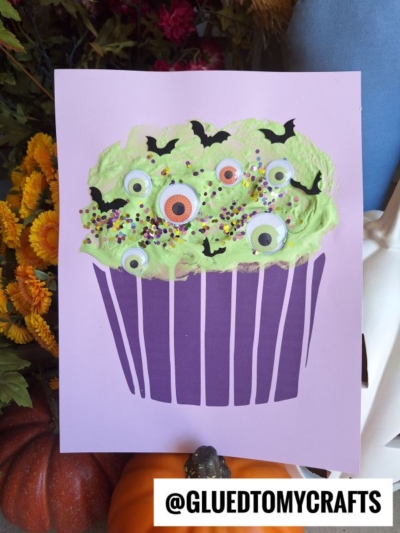 A puffy paint monster cupcake craft with green frosting, googly eyes, bat cutouts, and sprinkles on a lavender background, surrounded by fall decor. Text at the bottom reads @GLUEDTOMYCRAFTS.