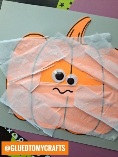 A pumpkin mummy drawing with googly eyes and a wavy mouth is partly wrapped in white tissue paper for a spooky effect. @GLUEDTOMYCRAFTS is written in a bright orange box at the bottom left.
