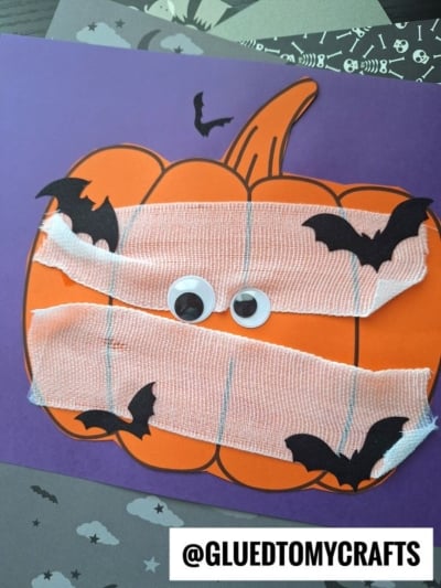 Paper pumpkin mummy decorated with white gauze, googly eyes, and black paper bats on a purple background. The handle @GLUEDTOMYCRAFTS is written at the bottom.