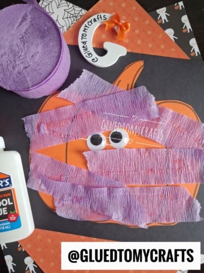 A pumpkin craft is covered with purple crepe paper and two googly eyes, transforming it into a playful pumpkin monster. Next to it are a bottle of glue, purple streamers, orange pennants, and a white keychain. @GLUEDTOMYCRAFTS is written at the bottom.