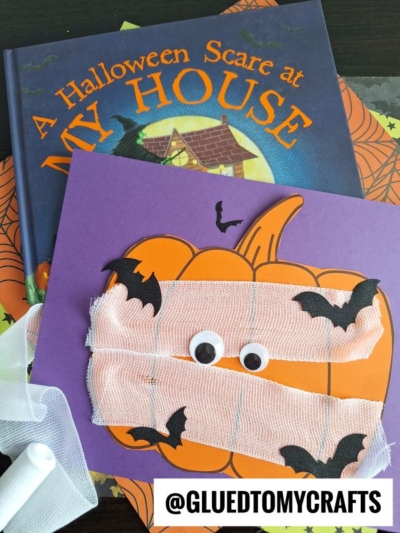 A children’s Halloween book and a handmade pumpkin mummy card decorated with googly eyes, white gauze, and black bat cutouts. The handle @GLUEDTOMYCRAFTS appears in the bottom right corner.