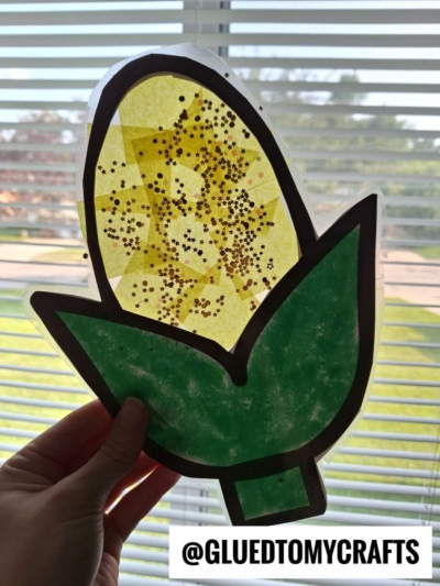 A hand displays a corn suncatcher craft made from colored paper and glitter in front of a window with blinds. Text at the bottom reads @GLUEDTOMYCRAFTS.