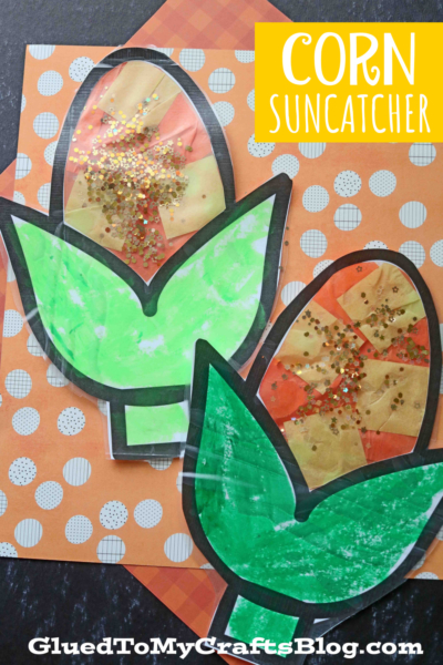 Two corn suncatcher crafts with green leaves, yellow and orange tissue paper, and gold glitter resemble stained glass corn, displayed on a polka-dot background. CORN SUNCATCHER and a craft blog name are written on the image.