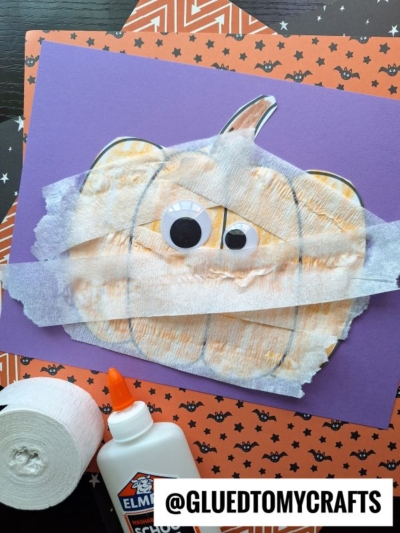 A pumpkin mummy drawing on purple paper is decorated with googly eyes and white tissue strips. Elmer’s glue, a tape roll, and patterned paper are nearby. Text at the bottom reads @GLUEDTOMYCRAFTS.