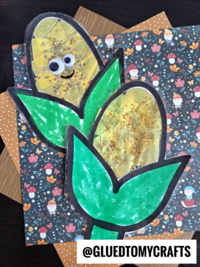 Two handmade corn crafts with green leaves and yellow textured kernels, one with googly eyes and a smile, placed on autumn-themed paper. These corn suncatchers add a playful touch. @GLUETOMYCRAFTS is written at the bottom.