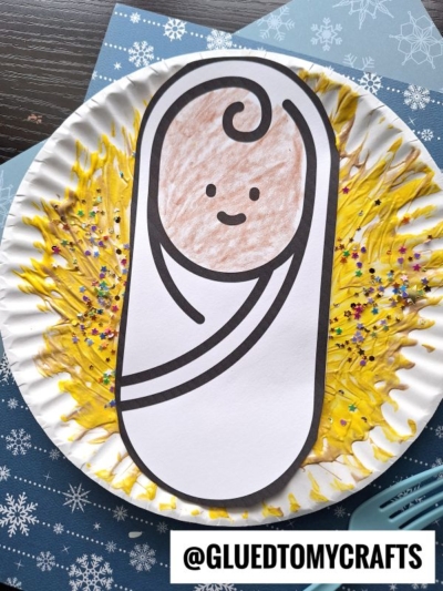 A Paper Plate Manger craft shows a swaddled baby with a smiling face in the center, surrounded by yellow paint and colorful glitter resembling straw. The image has a @GLUEDTOMYCRAFTS label at the bottom.