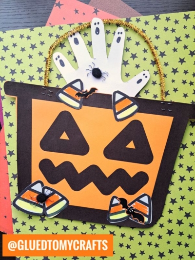 A Halloween craft featuring a candy bucket craft with a pumpkin bucket face, handprint ghosts, candy corn decorations, and fake spiders on star-patterned paper. @GLUEDTOMYCRAFTS is labeled in orange at the bottom left.