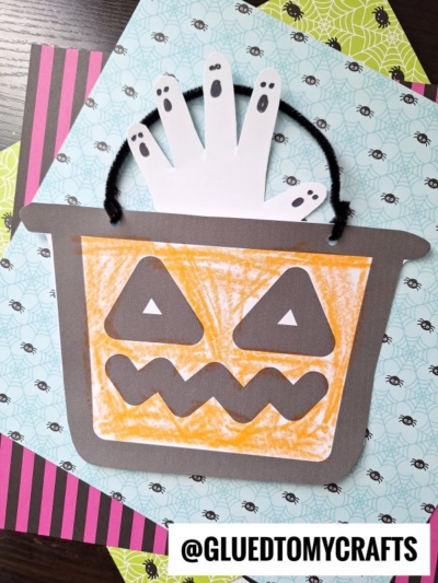 A Halloween candy bucket craft features a handprint ghost above an orange jack-o-lantern bucket, made with paper and crayons, on colorful spider-themed paper.