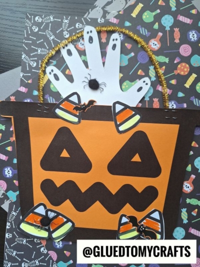 A Halloween candy bucket craft featuring a paper pumpkin with a jack-o-lantern face, decorated with a white handprint, candy corn cutouts, plastic spiders, and a gold pipe cleaner handle on a colorful spooky background.