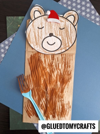 A paper bag sleeping bear wears a red Santa hat, its peaceful face and fur texture painted on using a blue-handled fork. The image is watermarked @GLUEDTOMYCRAFTS.