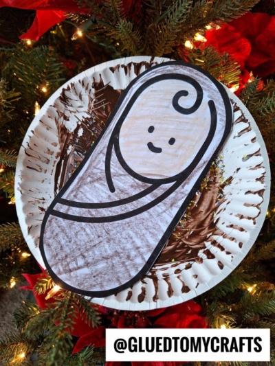 A Paper Plate Manger craft of baby Jesus with a smiling face, wrapped in a brown blanket, displayed on a Christmas tree with lights and red poinsettias. @gluedtomycrafts is written at the bottom.